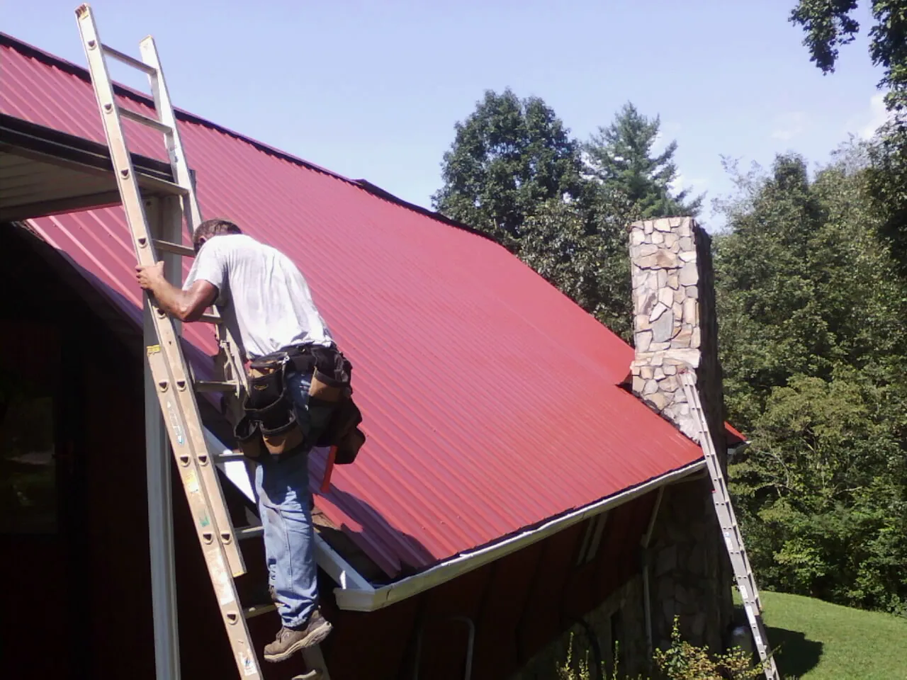 Roofer on a ladder beside a red metal roof and stone chimney — Soffit & Fascia Repair by Roofing Brothers in Blue Ridge